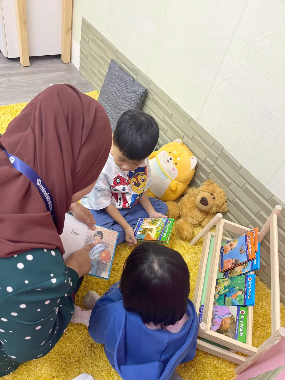 A teacher reading a picture book with two children on a yellow reading carpet